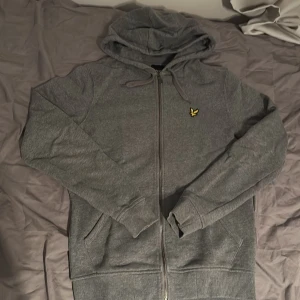 Grå hoodie från Lyle & Scott - Grå hoodie från Lyle & Scott med dragkedja, huva med snörning och klassisk gul logga på bröstet. Mjuk bomullskänsla, ribbade muddar och två fickor framtill. Perfekt för chill dagar eller när du vill ha en clean streetstyle.