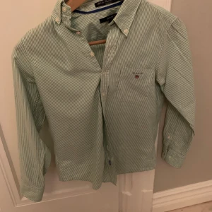 Grön- och vitrandig skjorta från Gant - Snygg grön- och vitrandig skjorta från Gant med klassisk button down-krage och bröstficka med broderad logga. Skjortan är i bomull och har långa ärmar samt knappar framtill. Perfekt för en clean och stilren look.