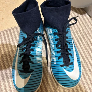 Nike MercurialX blå fotbollsskor - Snygga Nike MercurialX fotbollsskor i blått med svarta och vita detaljer. Skorna har högt skaft i stretchigt material, snörning och vit Nike-logga på sidan. Perfekta för dig som vill sticka ut på planen med stilren design och grym passform.