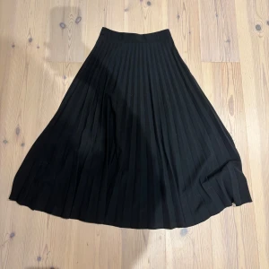 Svart plisserad långkjol - Svart långkjol med snygga plisserade veck som ger ett flowy och elegant intryck. Kjolen har hög midja och klassisk A-linjeform. Perfekt att styla med chunky sneakers eller boots för en cool look. Sömen vid dragkedjan har gått upp lite men syns inte när stängd och är förmodligen lätt att fixa