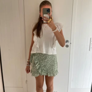 Ljusgrön blommig omlottkjol med volang - Superfin ljusgrön kort omlottkjol med vita blommor och volangkant. Kjolen har en söt knytning i sidan och är perfekt för vår och sommar. Lätt och luftig, passar till crop tops och sneakers.