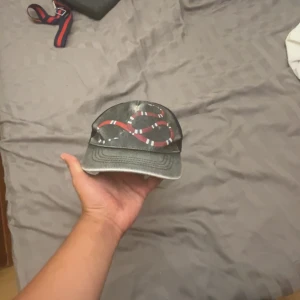 Svart keps från Gucci med orm - Säljer en svart keps från Gucci med en röd, vit och svart orm broderad framtill. Kepsen har justerbart band bak med rödblå rand och är tillverkad i mesh och bomull. Perfekt för dig som vill sticka ut med en lyxig accessoar. Säljer billigt p.g.a synlig användning 