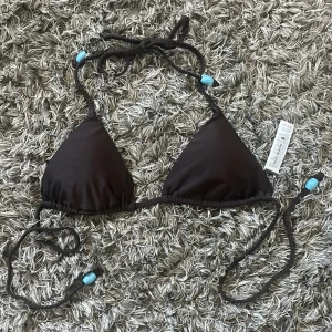 Brun bikiniöverdel BikBok XS - Mörkbrun trekantsbikini från BikBok storlek XS, med knyt i nacken och ryggen. Snygga ljusblå/turkosa pärlor på flätade snörena ger en extra detalj. Bikinin kan justeras längst snöret. Perfekt för stranden eller poolen under sommaren! 