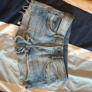Lågmidjade jeansshorts från H&M - Snygga ljusblå jeansshorts från H&M med låg midja och råa, fransiga kanter. Klassisk femficksmodell med knappar och dragkedja framtill. Perfekta för sommaren och ger en avslappnad vibe till din outfit.
