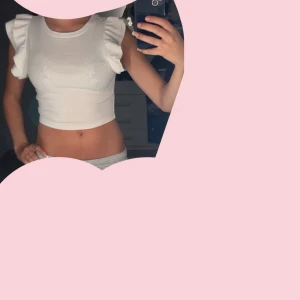 Vit croppad topp med volangärmar - Superfin vit croppad topp med volangdetaljer på axlarna. Ärmlös modell med rund halsringning och tight passform. Perfekt för dig som vill ha en söt och trendig look till sommaren.