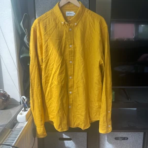 Gul långärmad skjorta från East West - Snygg gul skjorta från East West i storlek L. Skjortan har klassisk krage, knäppning framtill med vita knappar och långa ärmar. Perfekt för dig som vill sticka ut med färg och enkel stil. Materialet känns mjukt och passar bra till både jeans och chinos.