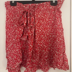 Jättefin röd blommig omlottkjol med volang - Superfin röd omlottkjol med vitt blommönster och volangkant. Går att knyta där fram, använd ett få antal gånger. Köparen står alltid för frakten!💞
