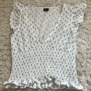 Blommig blus med volang från Gina Tricot - Supersöt blus från Gina Tricot i vitt med små blå blommor. Toppen har v-ringning, smockad midja och volangdetaljer vid ärmslut och nederkant. Lätt och luftig med korta volangärmar, perfekt för en somrig look. Köp för runt 300kr! Pris kan diskuteras 