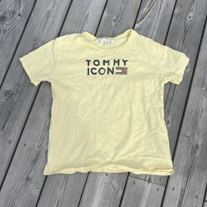 Gul t-shirt Tommy Hilfiger ICON - Gul t-shirt från Tommy Hilfiger med trycket 'TOMMY ICON' och en liten flaggdetalj i svart och rött framtill. Klassisk rund hals och korta ärmar. Tillverkad i mjuk bomull, perfekt för en avslappnad och cool stil.