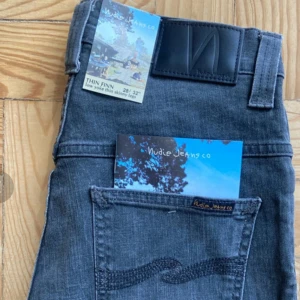 Nudie Jeans Thin Finn grå jeans - Säljer ett par gråa Nudie Jeans Thin Finn med skinny fit och klassisk broderad bakficka. Jeansen har svarta detaljer och är tillverkade i mjukt denimtyg. Perfekta för dig som gillar en smalare siluett och stilren look.