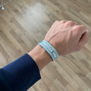 Blått armband Christian Dior J'Adior - Snyggt vävt armband från Christian Dior i ljusblått med vita detaljer och texten 'CHRISTIAN DIOR J'ADIOR' broderad. Armbandet har en enkel och stilren design med justerbar knytning och små fransar i ändarna.