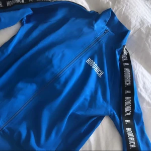 Blå zip tröja från Hoodrich - Snygg blå zip tröja från Hoodrich med dragkedja framtill och svarta band med vit logga längs ärmarna. Jackan har hög krage och sportig vibe, perfekt för dig som gillar streetwear. Materialet känns lätt och smidigt, och passformen är normal. 