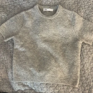 Grå stickad kortärmad tröja Zara - Enkel och stilren grå stickad tröja från Zara i storlek S. Tröjan har korta ärmar, rund halsringning och ribbade kanter vid ärmslut och nederkant. Perfekt för lager-på-lager och funkar året runt för en chill look.