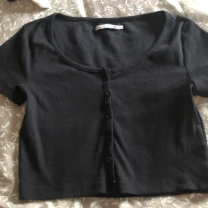 Svart croppad topp med knappar 157 - Svart croppad topp från 157 med korta ärmar och rund halsringning. Toppen har en ribbad struktur och knäppning med svarta knappar framtill. Perfekt att styla med höga jeans eller kjol för en chill look.