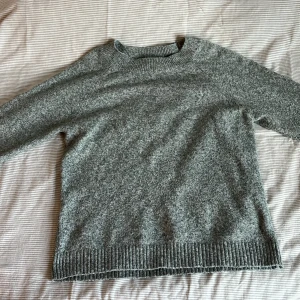 Grå grön stickad tröja från Vero Moda - Mysig grå grön stickad tröja från Vero Moda i storlek S. Tröjan har en klassisk rund halsringning, långa ärmar och ribbade muddar vid ärmslut och nederkant. Perfekt för lager-på-lager och chill höst- och vinterstil.