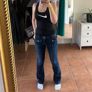 Mörkblå bootcut jeans med fickdetaljer - Snygga mörkblå jeans med bootcut passform och coola fickor framtill med lock och knappar. Jeansen har klassisk femficksdesign, kontrastsömmar och är tillverkade i stretchigt denim för skön passform. Perfekta för dig som gillar en retroinspirerad look. Helt oanvända 