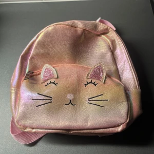Rosa ryggsäck med kattmotiv - Supersöt liten ryggsäck i skimrande rosa syntetmaterial med kattansikte och glittriga öron på framsidan. 📩 Skicka gärna ett meddelande vid frågor eller om du vill ha flera bilder! 🌸😊 📦 Jag skickar snabbt och packar alltid med omsorg!