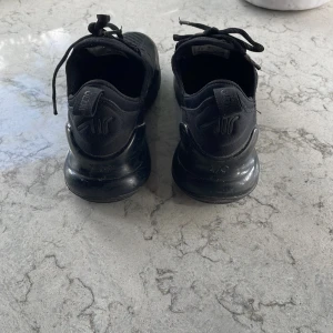 Svarta Nike Air Max 270 sneakers - Säljer ett par helsvarta Nike Air Max 270 sneakers med mesh-ovandel och synlig Air-enhet i hälen. Skorna har snörning, rund tå och en chunky sula som ger en sportig vibe. Perfekta för dig som gillar streetstyle och vill ha bekväma skor med snygg design.