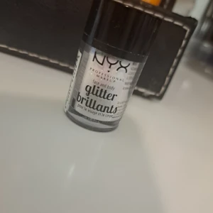 NYX Face & Body Glitter Silver - Glittrigt lösglitter från NYX i färgen silver, perfekt för att skapa extra glow på både ansikte och kropp. Kommer i en liten burk med svart lock och är lätt att applicera för en riktigt festlig look. Materialet är syntetiskt glitter.
