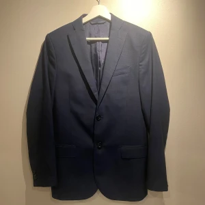 Mörkblå kavaj från J.Lindeberg - Snygg mörkblå kavaj från J.Lindeberg med klassisk passform. Kavajen har två knappar framtill, fyra knappar vid ärmslut och diskreta fickor. Storlek S och passar S!