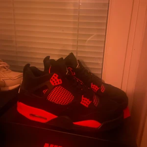 Nike Air Jordan 4 Svart/Röd - Nike Air Jordan 4 sneakers i svart mocka med röda detaljer och meshpaneler. Klassisk siluett med snörning, synlig Air-enhet i sulan och Jumpman-logga på plösen. Snygg kontrast mellan svart och rött gör dem till ett statement på fötterna. Slängd boxen då jag köpte den för länge sen och använt skorna max 5ggr.