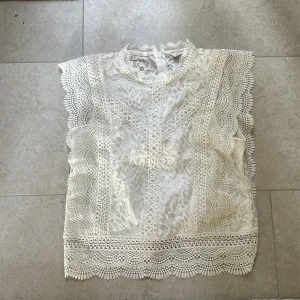 Vit spetsblus från Only - Superfin vit blus i spets med högt halsringning och vackra broderade detaljer. Blusen är ärmlös med breda spetskanter och har ett romantiskt, feminint uttryck. Perfekt att styla med jeans eller kjol för en trendig look.