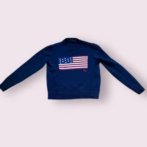 Mörkblå stickad tröja Polo Ralph Lauren - Säljer en mörkblå stickad tröja från Polo Ralph Lauren med amerikansk flagga och RL-broderi på bröstet. Tröjan har ribbade muddar och lång ärm, perfekt för dig som gillar klassisk preppy stil., storlek L men krympt i tvätten så den sitter som en S