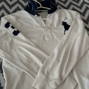 Vit pikétröja från Ralph Lauren - Vit långärmad pikétröja från Ralph Lauren med klassisk krage i blått med gula ränder. Stor marinblå logga på bröstet och siffran 3 på ena ärmen. Tröjan har ribbade muddar och knäppning vid halsen. Perfekt för en sportig och preppy look.
