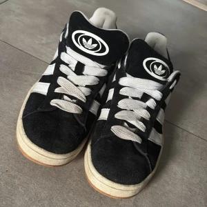 Adidas Campus svarta sneakers mocka - Svarta Adidas Campus sneakers i mocka med vita ränder och vit sula. Klassisk retrostil med rund tå och snörning. Ikonisk Adidas-logga på plösen och extra svarta skosnören medföljer. Perfekta för dig som gillar streetwear och tidlös design. De är använda max 3 gånger och säljs pga att de inte passar