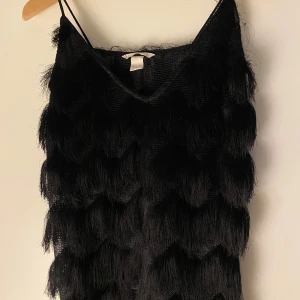 Svart fransig linnetopp från H&M - Svart linnetopp från H&M med tunna axelband och flera lager långa fransar som ger en riktigt cool och iögonfallande look. Toppen har en lätt transparent meshbas och är perfekt för dig som vill sticka ut med en unik stil🥂
