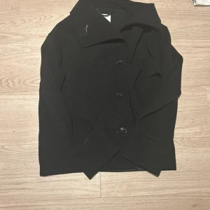 Svart kort kappa med klassisk krage - Stilren svart kort kappa med klassisk krage och tre knappar framtill. Jackan har en rak passform och långa ärmar, perfekt för dig som gillar en clean och tidlös look. Passar lika bra till jeans som till kostymbyxor.