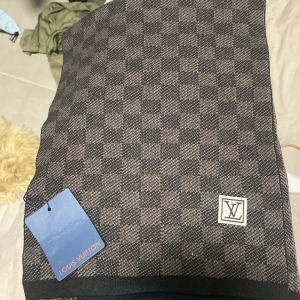 Rutig halsduk från Louis Vuitton - Snygg rutig halsduk från Louis Vuitton i mörkbruna och grå toner. Halsduken har klassiskt LV-märke och är tillverkad i en lyxig mix av 90% ull och 10% kashmir. Perfekt accessoar för att lyfta din outfit med exklusiv känsla.