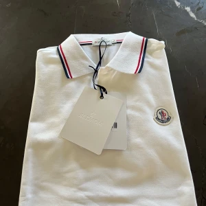 Vit pikétröja från Moncler - Snygg vit pikétröja från Moncler med klassisk krage och röd-blå ränder på krage och ärmslut. Tröjan har Moncler-logga broderad på bröstet och är tillverkad i bomull för en fräsch och stilren look. Perfekt för dig som gillar stilrena och sportiga vibes. Små i storlek så denna passar bättre i M. Pris kan diskuteras vid snabb affär 