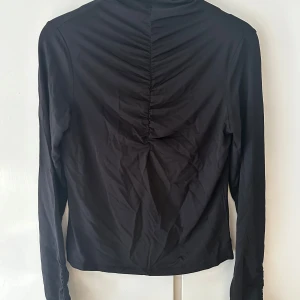 Svart långärmad topp från Indiska - Svart långärmad topp från Indiska i stretchigt material. Snygg rynkdetalj längs ryggen och ärmarna ger en cool och modern look. Perfekt att styla med jeans eller kjol för en trendig outfit.