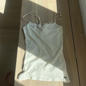 Vit ribbad linnetopp från Hollister - Supersöt vit ribbad linnetopp från Hollister med tunna justerbara axelband och spetsdetaljer framtill. Toppen har en v-ringad urringning med broderade blommor och är perfekt för varma dagar. Enkel att matcha med jeans eller kjol.