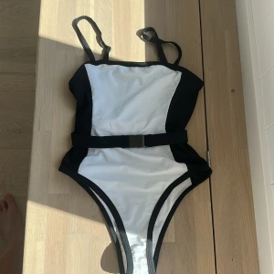 Svartvit baddräkt från NA-KD x Hannalicious - Snygg svartvit baddräkt från NA-KD x Hannalicious med justerbara axelband och ett coolt bälte i midjan. Hög benskärning och stilren design som ger en sportig vibe. Perfekt för stranden eller poolen!