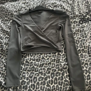 Grå omlottopp från Gina Tricot - Snygg grå/ grön croppad omlottopp från Gina Tricot med lång ärm och djup v-ringning. Toppens stilrena design gör den lätt att matcha och den har en tight passform som framhäver siluetten.