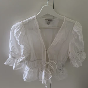 Vit blus med knyt från AWARE - Supersöt vit blus från AWARE i storlek XS. Blusen har puffiga korta ärmar, broderade spetsdetaljer och volangkant. Den är croppad och knyts framtill för en romantisk vibe. Perfekt till jeans eller kjol för en fräsch look.