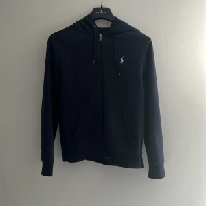 Marinblå tröja från Polo Ralph Lauren - Snygg marinblå hoodie från Polo Ralph Lauren med dragkedja och klassisk vit logga på bröstet. Nypris 1499kr. Använd under 1 år. Storlek small. 