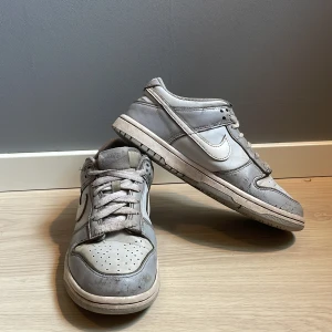 Nike Dunk Low i grått skinn - Nike Dunk Low sneakers i ljusgrått skinn med vita detaljer och klassisk swoosh på sidorna. Skorna har snörning, perforerad tå och en platt sula. Perfekta för dig som gillar en clean och tidlös stil.skostorleken är 38.