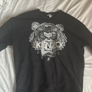 Svart Kenzo sweatshirt med tigerbrodyr - Svart sweatshirt från Kenzo med ikonisk silverbroderad tiger och logga på bröstet. Tröjan har rund hals, långa ärmar. Perfekt plagg för dig som vill sticka ut med stil. Inte den tjockaste materialet, och använt många gånger. Passar lite tajt på medium, alltså passar den small också!