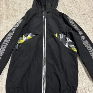 Svart Montreal vindjacka med tryck - Svart vindjacka från Montreal med huva och hel dragkedja framtill. Jackan har coola grafiska detaljer i vitt och gult på sidorna och ryggen samt stora Montreal-loggor längs båda ärmarna. Perfekt för dig som gillar streetstyle och sportig vibe.