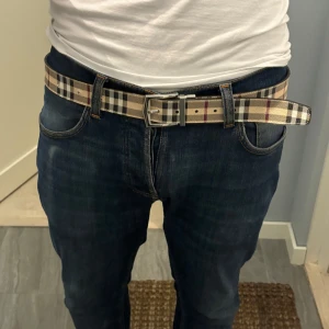 Rutig Burberry-bälte i skinn - Snyggt bälte från Burberry med klassiskt rutigt mönster i beige, svart, vitt och rött. Bältet har en fyrkantig metallspänne och är gjort i skinn med textil på ovansidan. Vet inte om de är äkta eller inte.