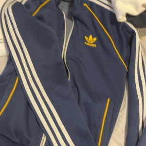 Blå Adidas bomberjacka med gula detaljer - Snygg blå bomberjacka från Adidas med klassiska vita ränder längs ärmarna och gula detaljer vid fickor och axlar. Jackan har dragkedja framtill, Adidas-logga på bröstet och ribbade muddar. Perfekt för dig som gillar sportig och retro stil.