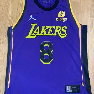 Lakers Bryant basketlinne lila-gult - Snyggt Lakers basketlinne i lila med gula detaljer och nummer 8, Bryant på ryggen. Klassisk NBA-design med Jordan-logga och Bibigo-märke på bröstet. Ärmlös modell i lätt polyester, perfekt för basket eller streetwear.