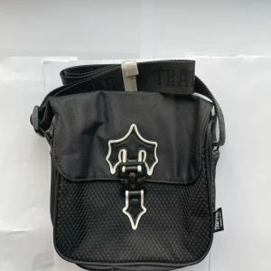Svart axelväska från Trapstar - Cool svart axelväska från Trapstar med justerbar rem och unik vitdetaljerad spänne framtill. Väskan är tillverkad i slitstark syntet och har ett meshmönstrat fack på framsidan. Perfekt för dig som gillar alternativ stil och vill sticka ut.