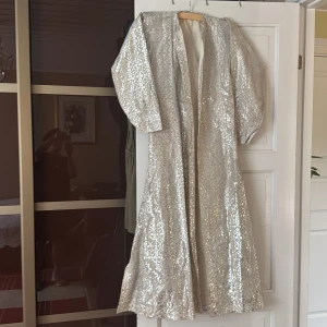 Lång glittrig silverklänning - Säljer en maxilång klänning i glittrigt silvertyg med vackert mönster. Klänningen har vida ärmar och en rak, elegant passform. Perfekt för dig som vill sticka ut med en riktigt glamorös look.