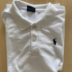Vit pikétröja från Polo Ralph Lauren - Passar runt 160! Klassisk vit pikétröja från Polo Ralph Lauren i 100% bomull. Tröjan har krage, knäppning med två knappar och den ikoniska broderade loggan i mörkblått på bröstet. Perfekt för en clean och stilren look. Vit eller svart för 250 kr båda för 400