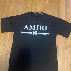 Svart AMIRI t-shirt med logga - Svart t-shirt från AMIRI med stor vit logga och grafisk detalj framtill. Klassisk rund halsringning och korta ärmar. Tillverkad i mjuk bomull för en skön och avslappnad känsla. Perfekt för dig som gillar streetwear och vill sticka ut med en exklusiv look.