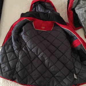 Röd vinterjacka Northbrook Sports - Röd vinterjacka från Northbrook Sports med quiltat svart foder och praktisk huva. Jackan har flera fickor med dragkedja och tryckknappar, samt snygga detaljer i svart. Perfekt för kalla dagar och riktigt skön passform.